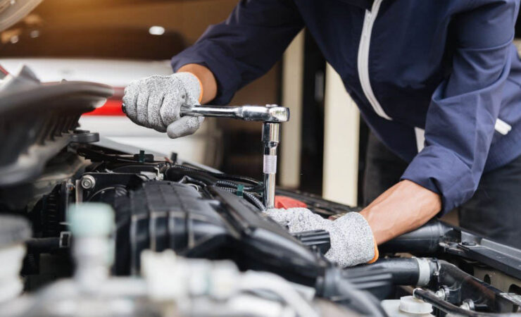 A mechanic, wearing a grease-stained uniform and safety glasses, leans over a car engine and uses a socket wrench to tighten a bolt.