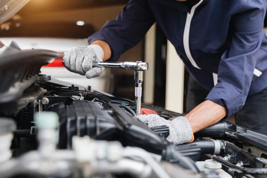 A mechanic, wearing a grease-stained uniform and safety glasses, leans over a car engine and uses a socket wrench to tighten a bolt.