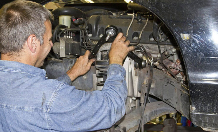 A mechanic, wearing a grease-stained uniform and work gloves, uses a tire iron to carefully remove a damaged tire from a wheel rim.