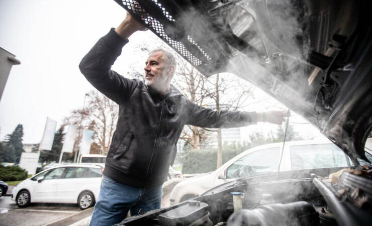 A silver car parked on the side of the road with smoke billowing from under the hood.