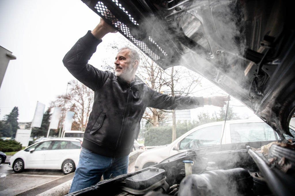 A silver car parked on the side of the road with smoke billowing from under the hood.