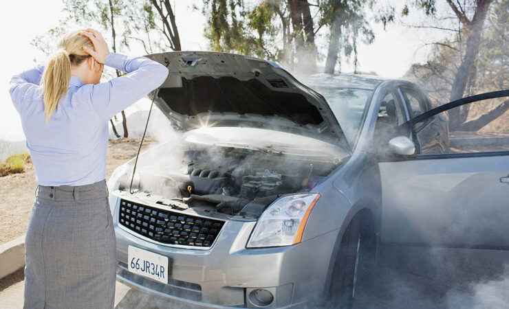 A distressed woman in a casual outfit, holding a phone, stands beside her car on the side of a road, smoke billowing from under the hood.