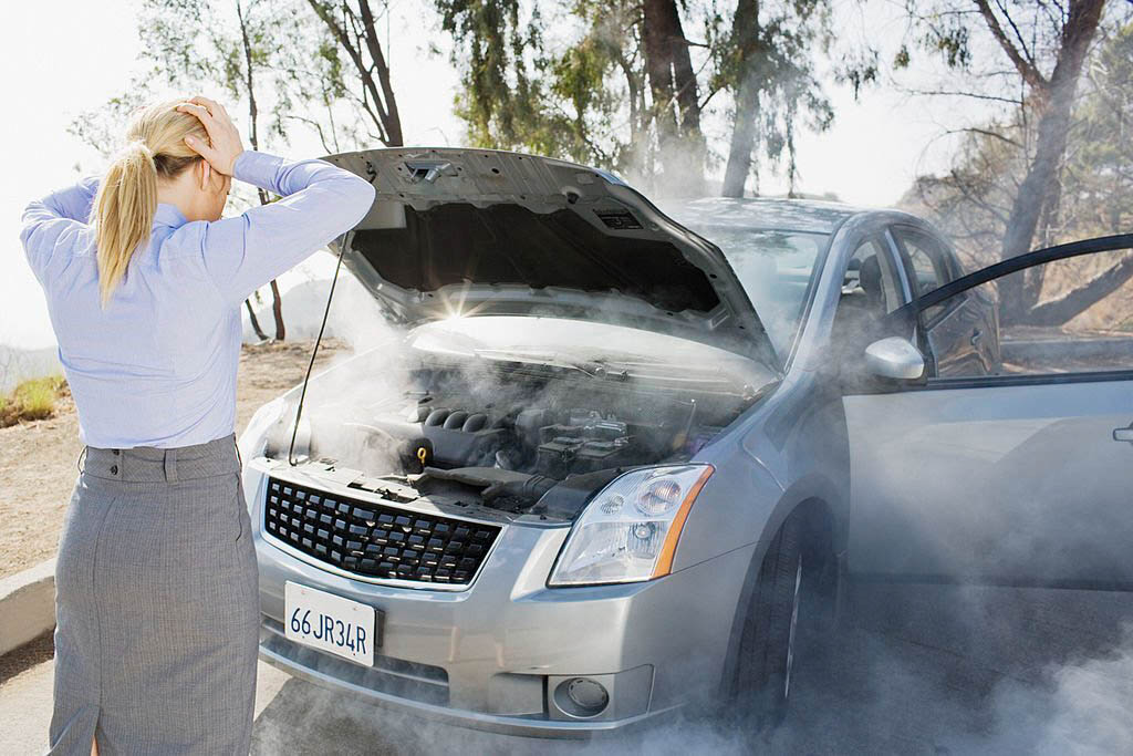 A distressed woman in a casual outfit, holding a phone, stands beside her car on the side of a road, smoke billowing from under the hood.