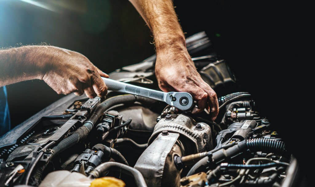 A mechanic, wearing a grease-stained uniform and safety glasses, leans over a car engine and uses a socket wrench to tighten a bolt.