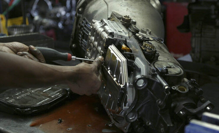 A mechanic, wearing a grease-stained uniform, uses a Phillips-head screwdriver to tighten a screw on a car's engine.