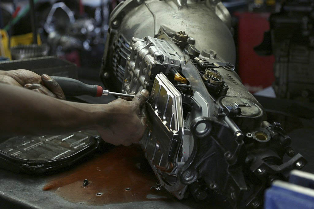 A mechanic, wearing a grease-stained uniform, uses a Phillips-head screwdriver to tighten a screw on a car's engine.