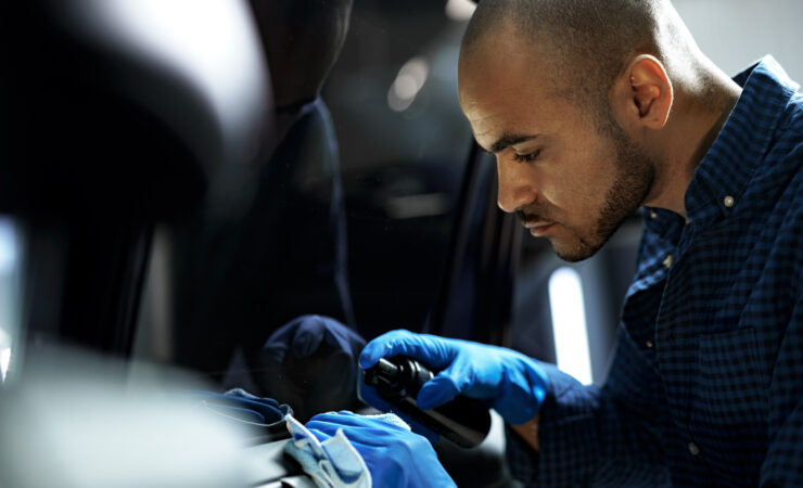 A focused African American car detailer in a protective suit carefully applies nano-coating to a car's hood using a soft applicator pad.