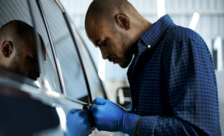 A focused African American car service worker in a protective mask and gloves carefully applies a nano-ceramic coating to a black car's hood.