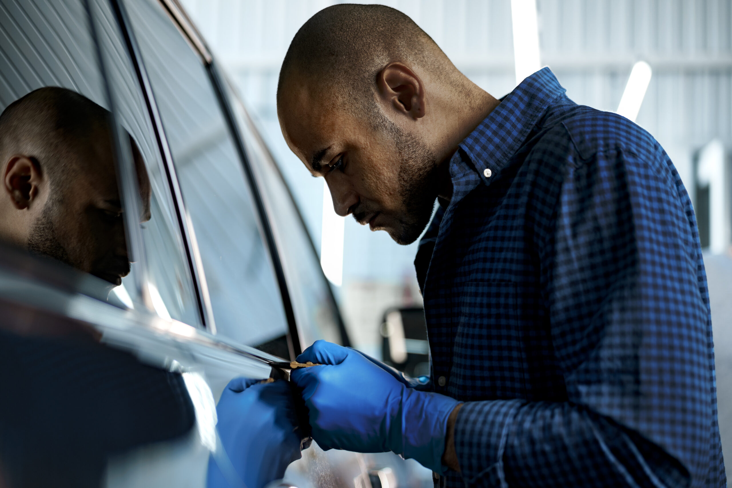 A focused African American car service worker in a protective mask and gloves carefully applies a nano-ceramic coating to a black car's hood.