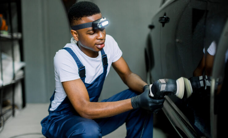 A professional detailer, wearing protective gear, carefully polishes a car's hood with a dual-action polisher and microfiber pad.