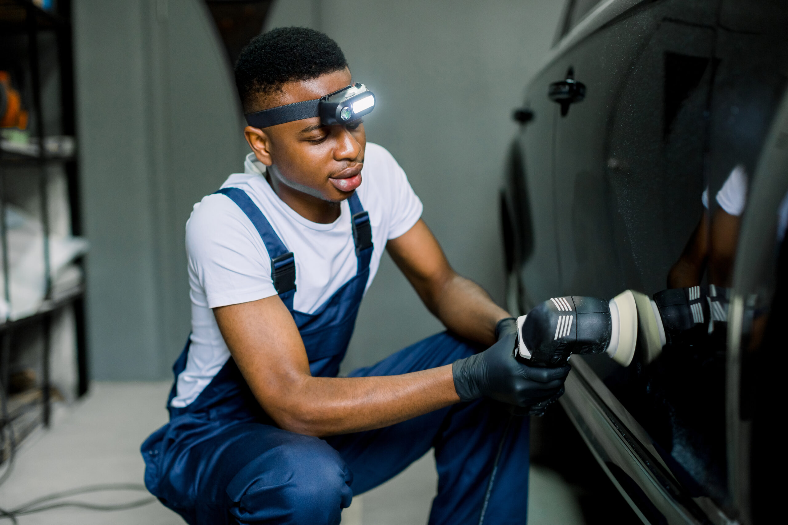 A professional detailer, wearing protective gear, carefully polishes a car's hood with a dual-action polisher and microfiber pad.