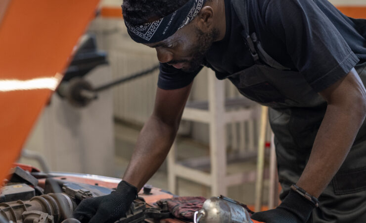 A Black car mechanic in a blue jumpsuit, with tools in hand, leans over the open hood of a car in a well-lit auto repair shop.v