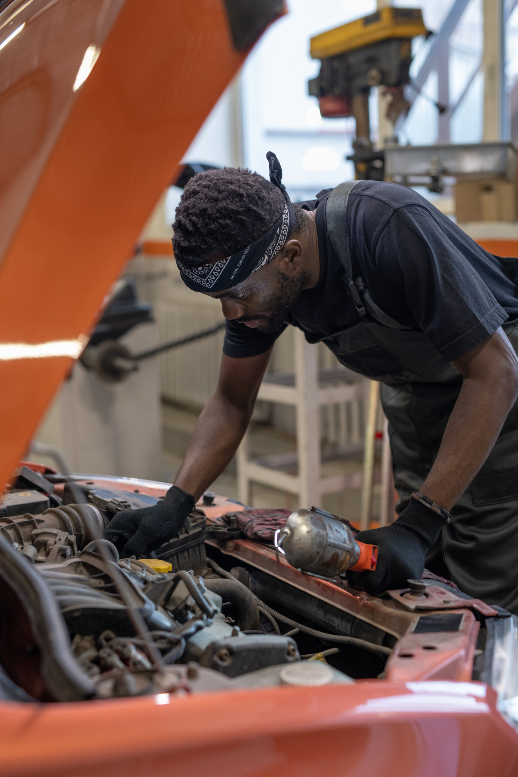 A Black car mechanic in a blue jumpsuit, with tools in hand, leans over the open hood of a car in a well-lit auto repair shop.v