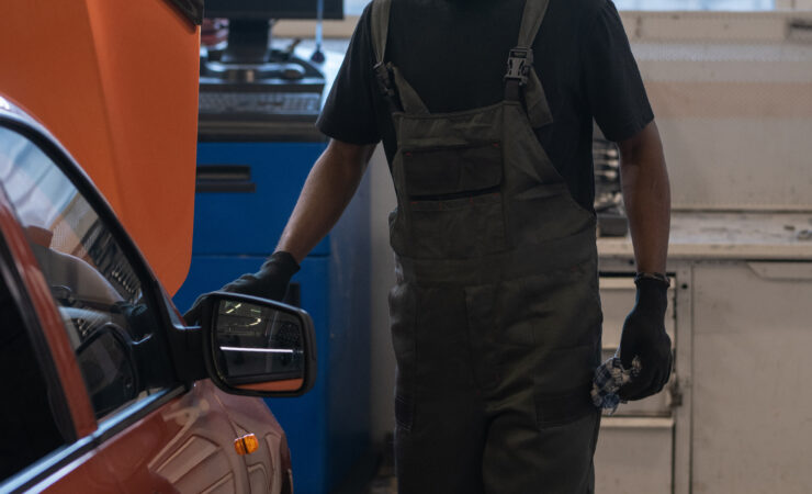 A focused Black mechanic, wearing a blue jumpsuit and safety glasses, tightens a bolt under the hood of a car.