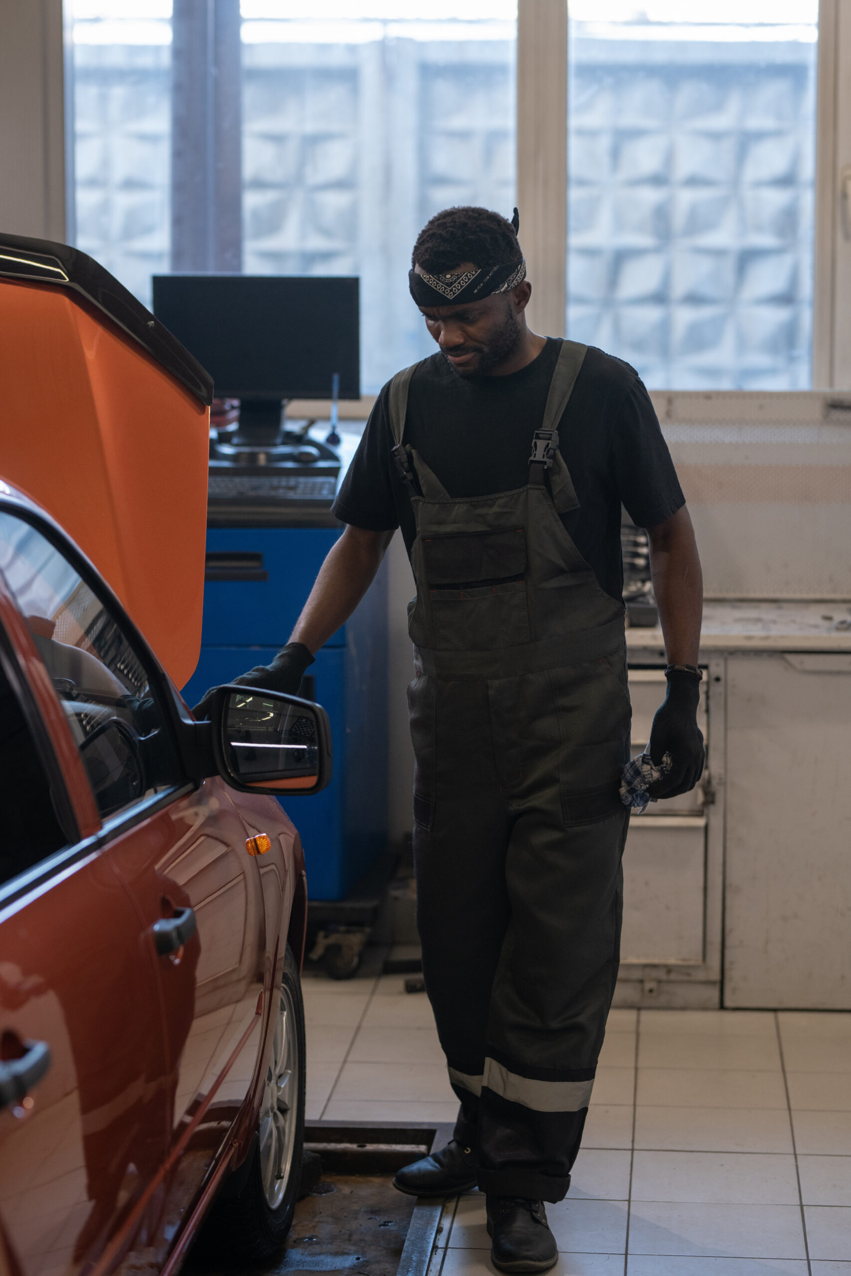 A focused Black mechanic, wearing a blue jumpsuit and safety glasses, tightens a bolt under the hood of a car.