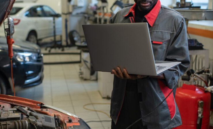 A Black mechanic in a garage, wearing a blue jumpsuit and safety glasses, uses a laptop computer to diagnose a car's issue.