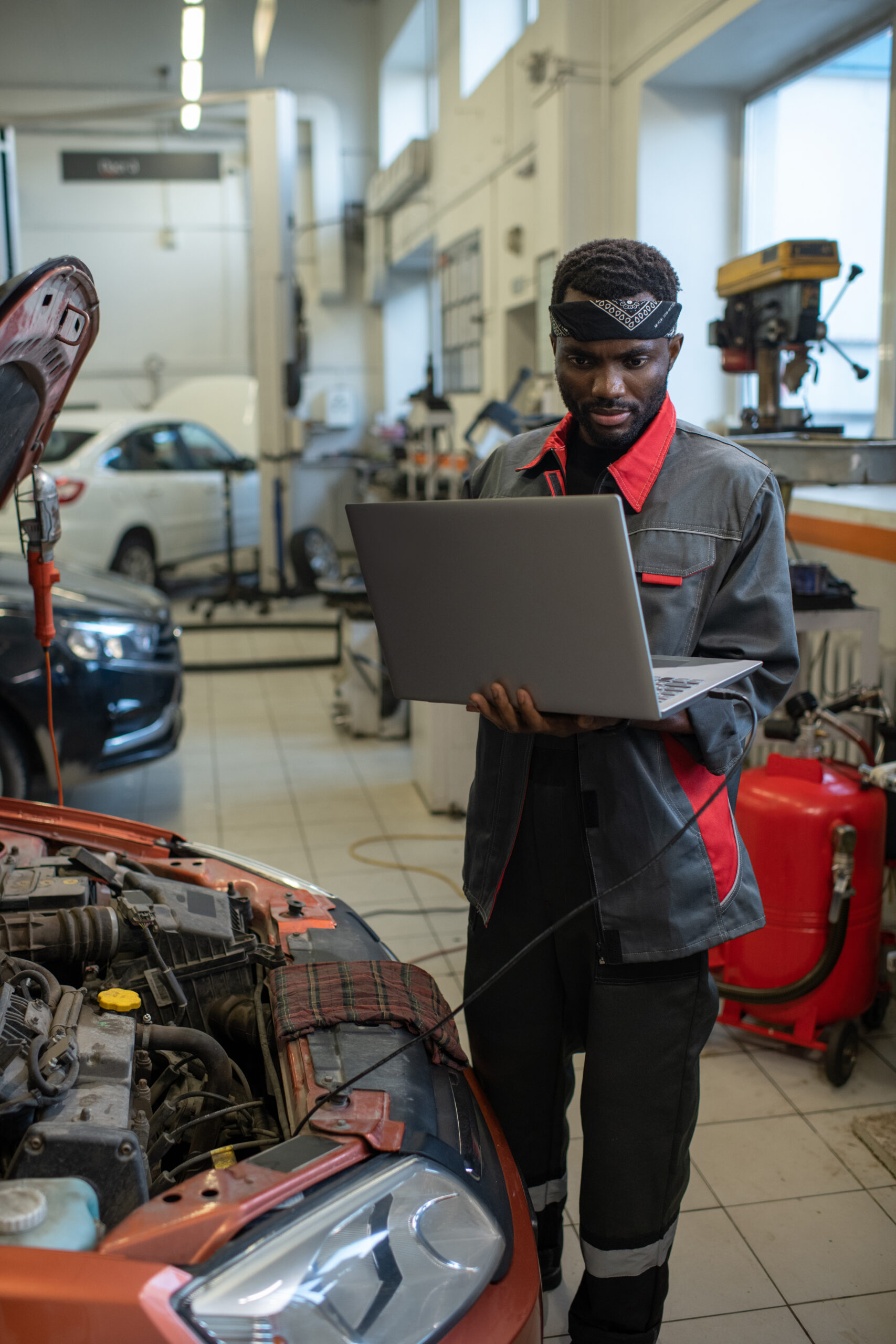 A Black mechanic in a garage, wearing a blue jumpsuit and safety glasses, uses a laptop computer to diagnose a car's issue.