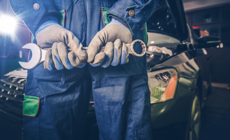 A car mechanic, dressed in a blue jumpsuit and holding a wrench, stands confidently in front of a tool chest in a well-lit garage.