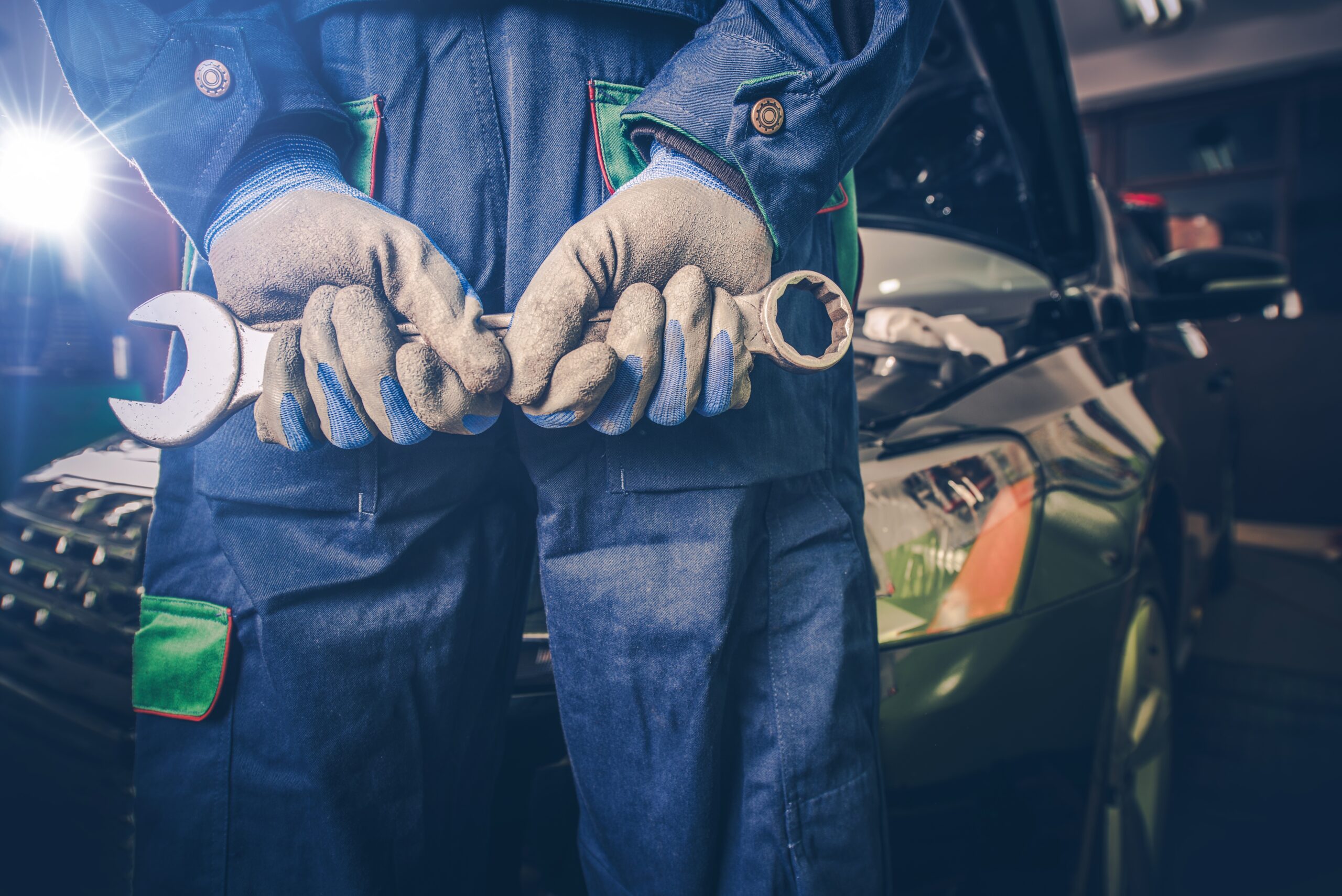 A car mechanic, dressed in a blue jumpsuit and holding a wrench, stands confidently in front of a tool chest in a well-lit garage.