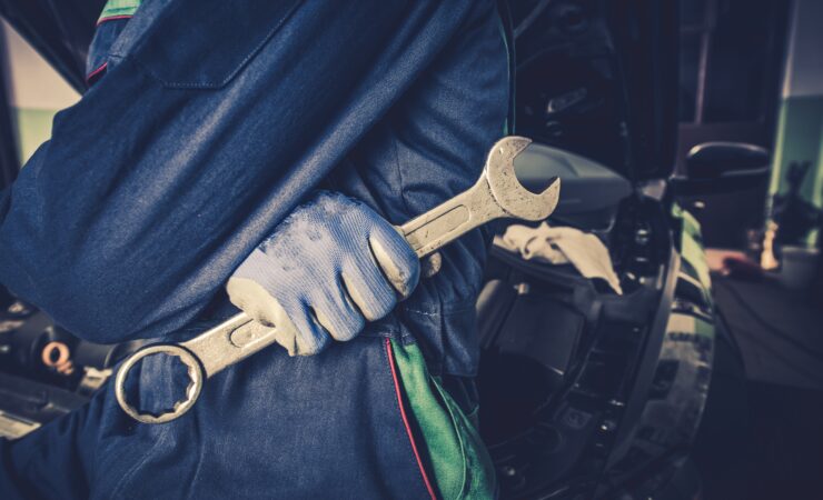 A focused car mechanic, wearing a blue jumpsuit and work boots, tightens a bolt on a car engine using a wrench.