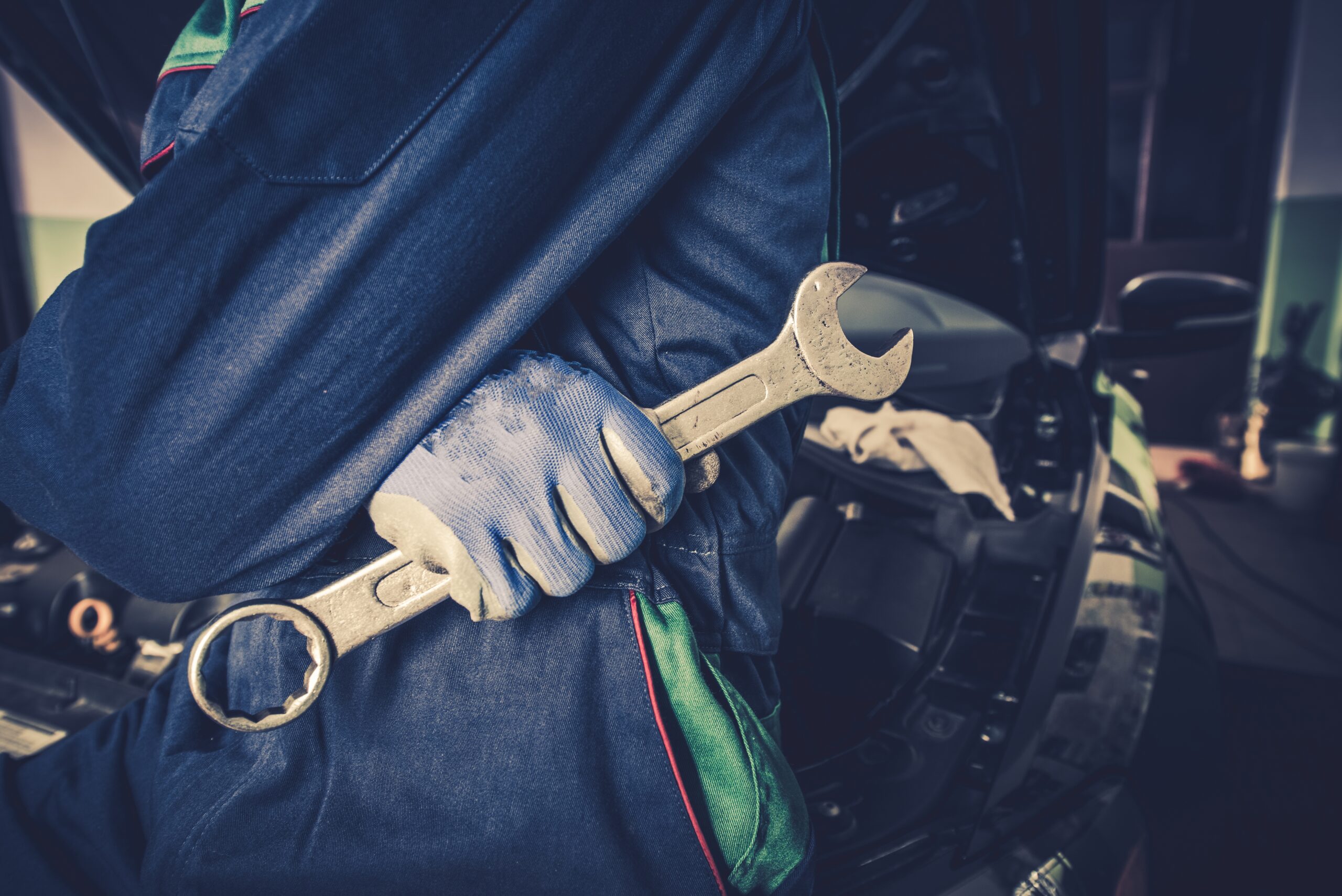 A focused car mechanic, wearing a blue jumpsuit and work boots, tightens a bolt on a car engine using a wrench.