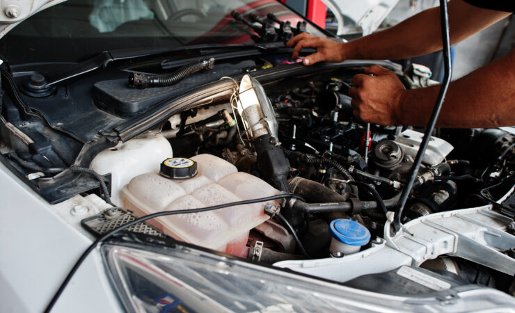 A mechanic in a blue jumpsuit lies under a car on a lift, using a wrench to tighten a bolt on the undercarriage.