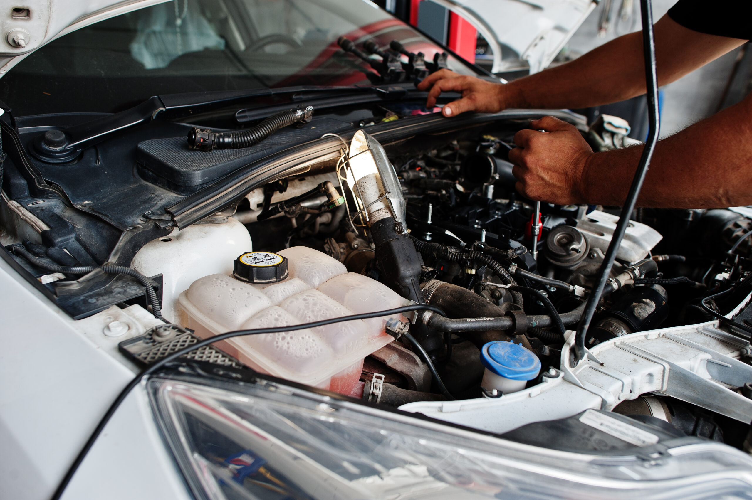 A mechanic in a blue jumpsuit lies under a car on a lift, using a wrench to tighten a bolt on the undercarriage.