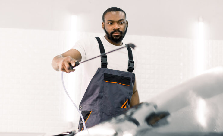 A young African man, wearing a car wash uniform, diligently cleans the windshield of a luxury car with a high-pressure water sprayer.