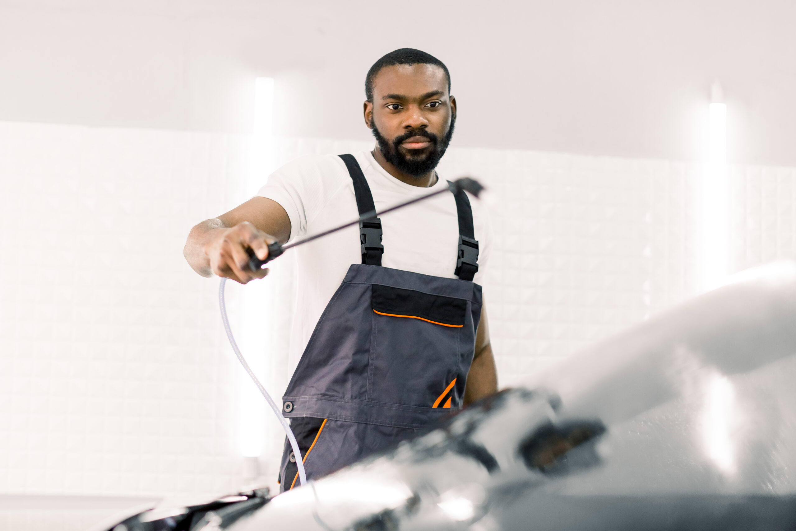 A young African man, wearing a car wash uniform, diligently cleans the windshield of a luxury car with a high-pressure water sprayer.