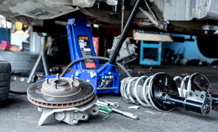 A mechanic, wearing safety glasses and gloves, inspects the brake calipers and rotors of a raised car.