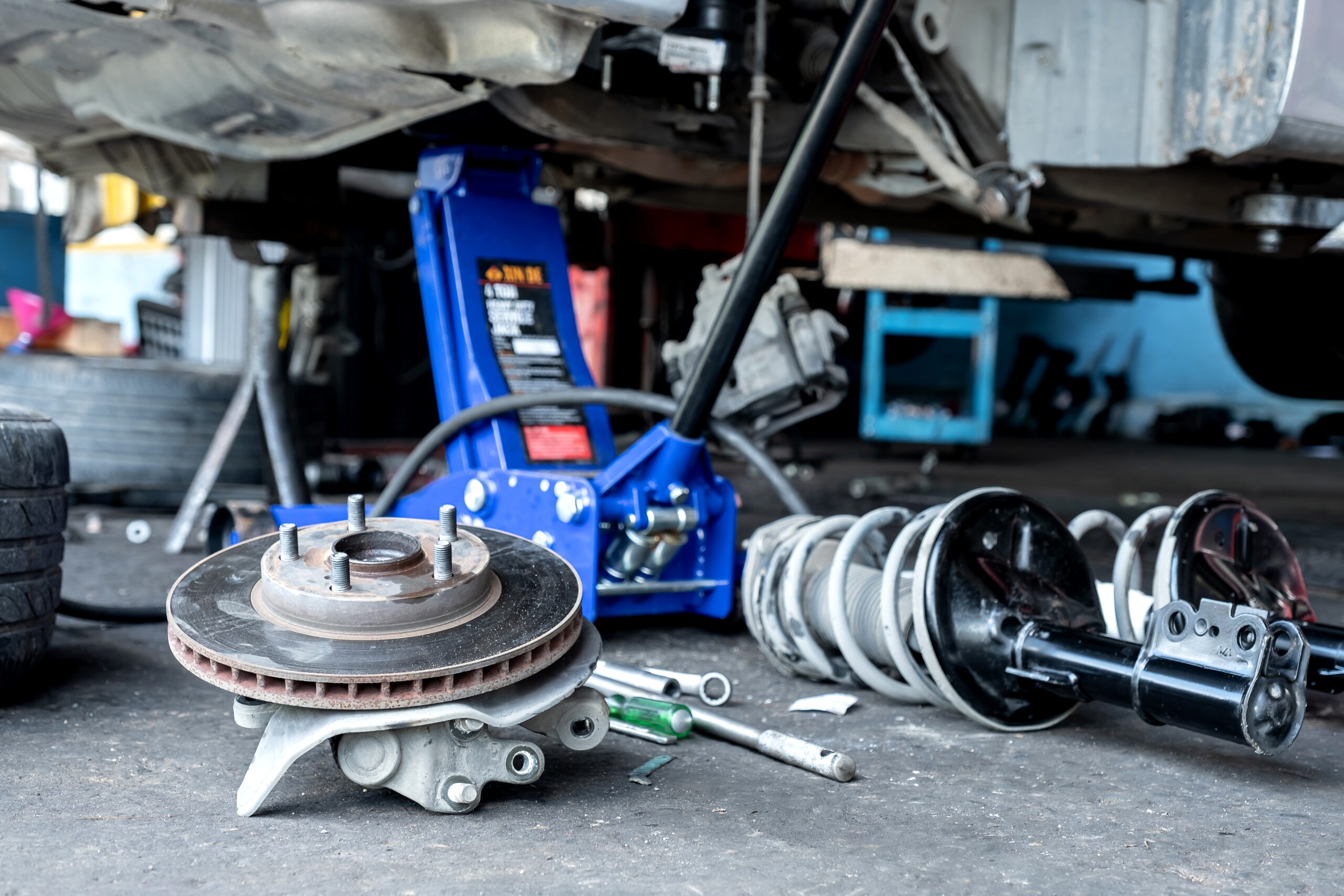 A mechanic, wearing safety glasses and gloves, inspects the brake calipers and rotors of a raised car.