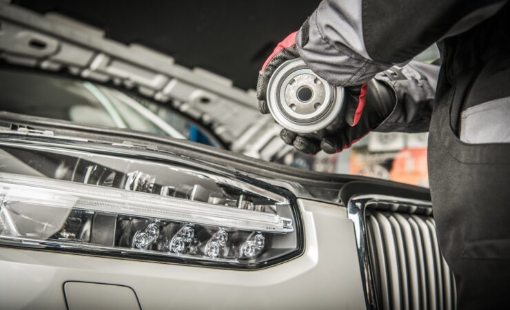 A mechanic's hand, wearing a black nitrile glove, reaches into a car's engine bay to unscrew a cylindrical oil filter.