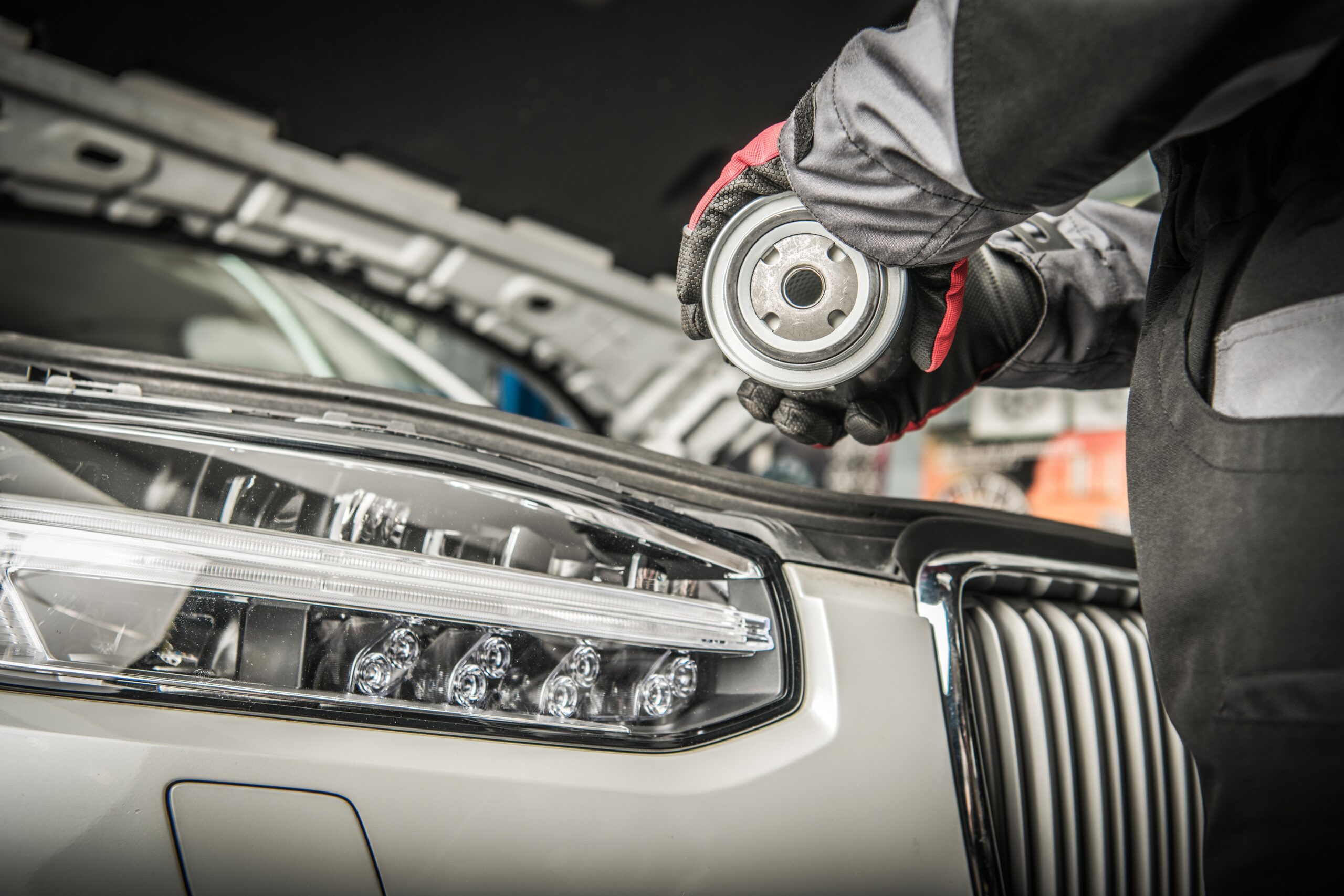 A mechanic's hand, wearing a black nitrile glove, reaches into a car's engine bay to unscrew a cylindrical oil filter.