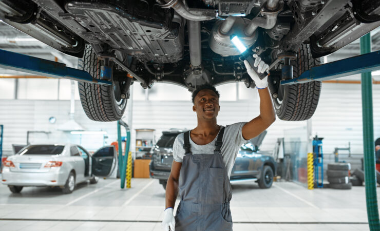 A male mechanic, wearing a blue jumpsuit and safety glasses, leans over the wheel well of a raised car, inspecting the suspension components.