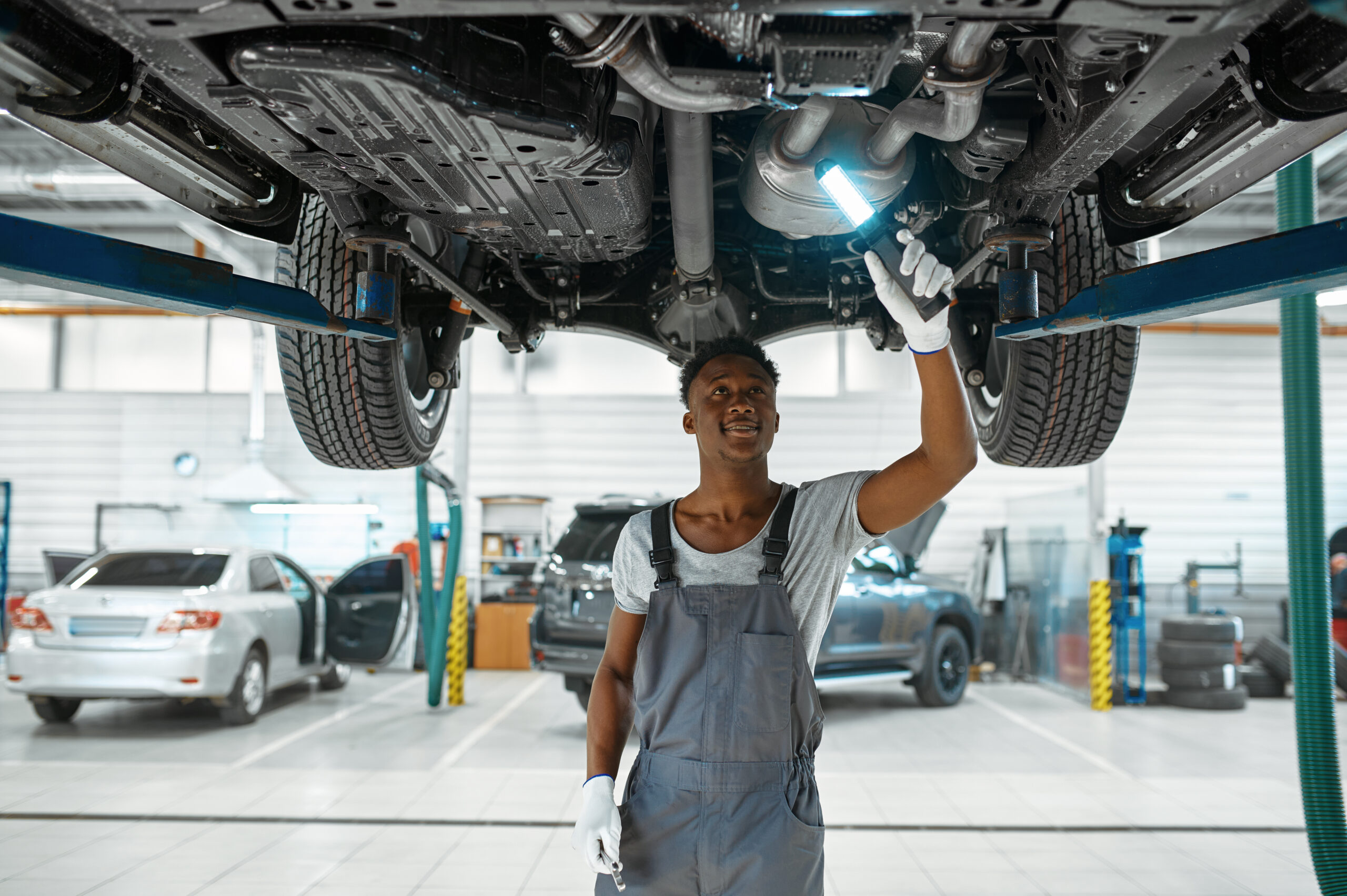 A male mechanic, wearing a blue jumpsuit and safety glasses, leans over the wheel well of a raised car, inspecting the suspension components.