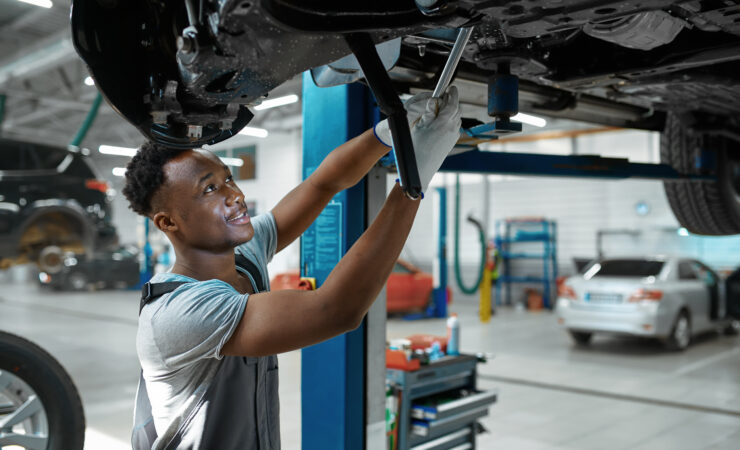 A male mechanic, wearing a blue jumpsuit and safety glasses, inspects a car's suspension components under the wheel well.