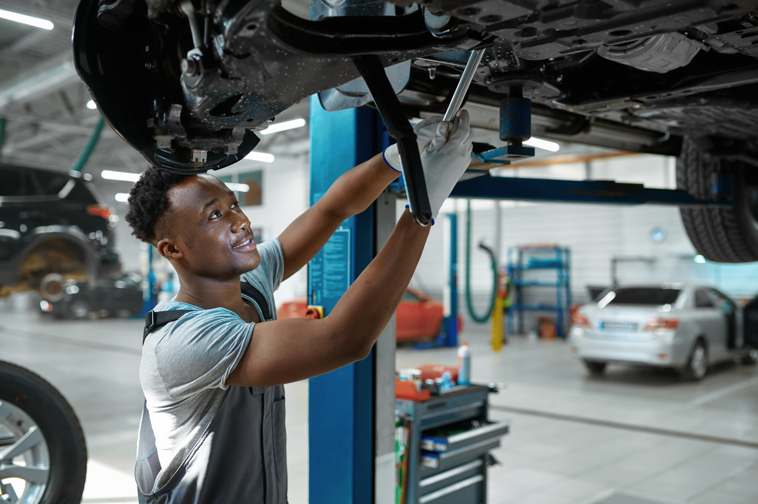 A male mechanic, wearing a blue jumpsuit and safety glasses, inspects a car's suspension components under the wheel well.