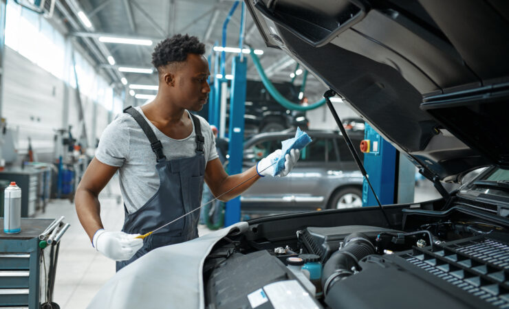 A male mechanic, wearing a blue jumpsuit and gloves, holds a dipstick up to the light, carefully checking the oil level in a car engine.