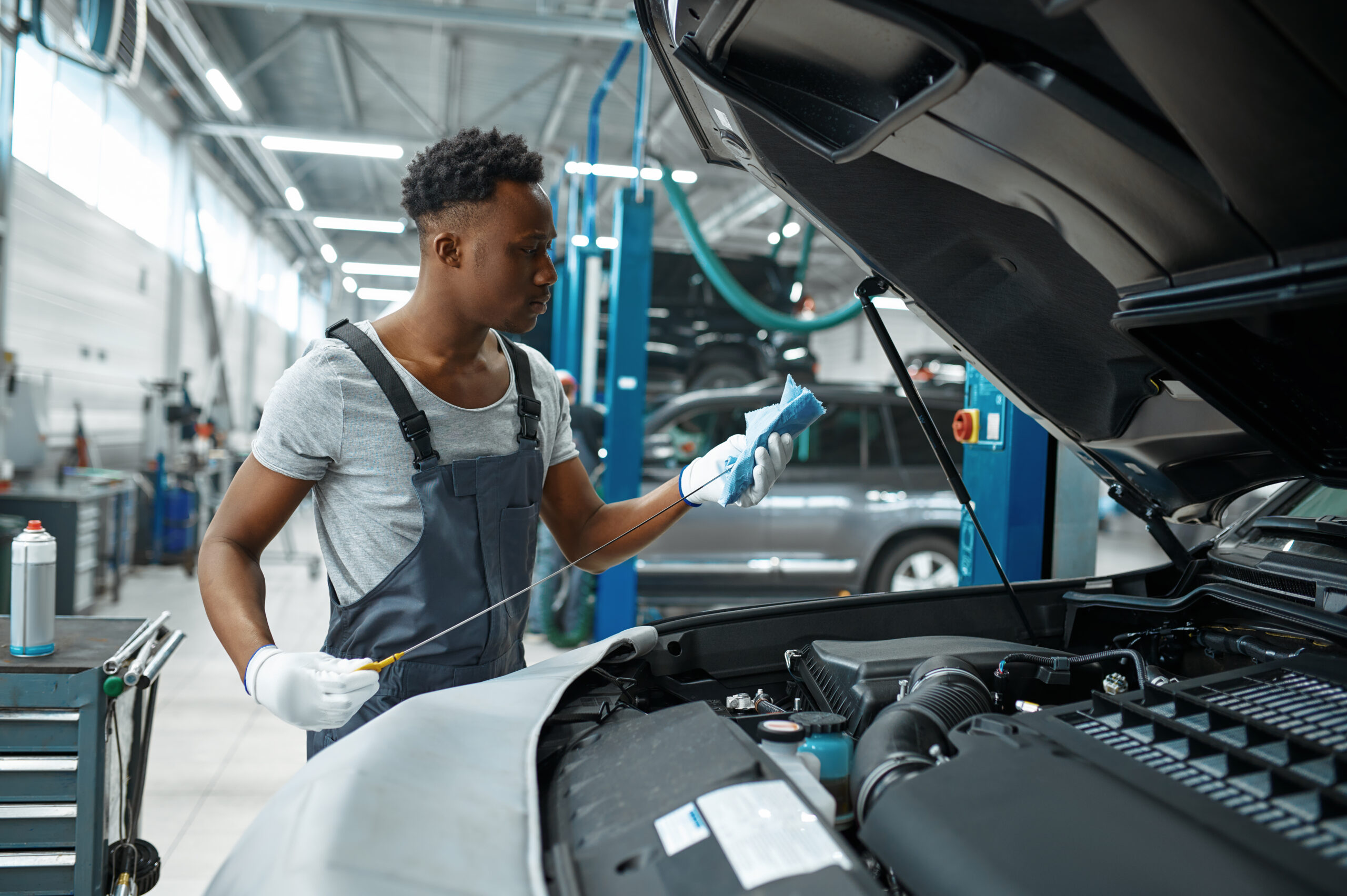 A male mechanic, wearing a blue jumpsuit and gloves, holds a dipstick up to the light, carefully checking the oil level in a car engine.