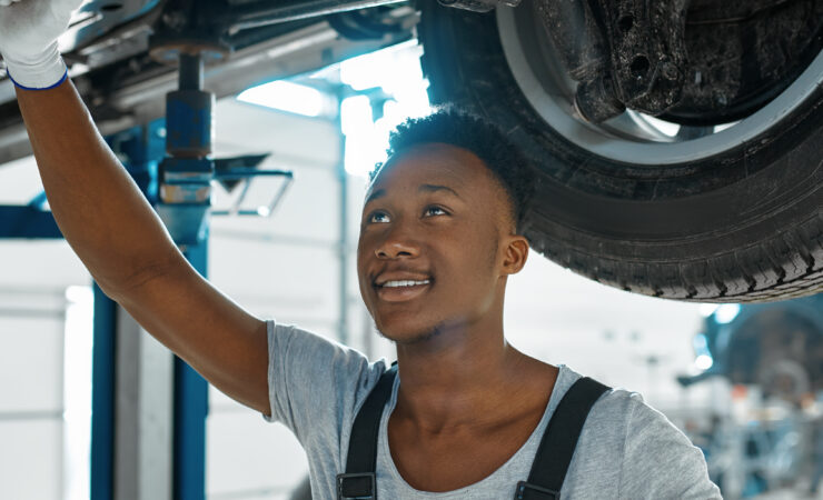 A male mechanic, wearing coveralls and safety glasses, carefully drains used motor oil from a car's engine into a collection pan.