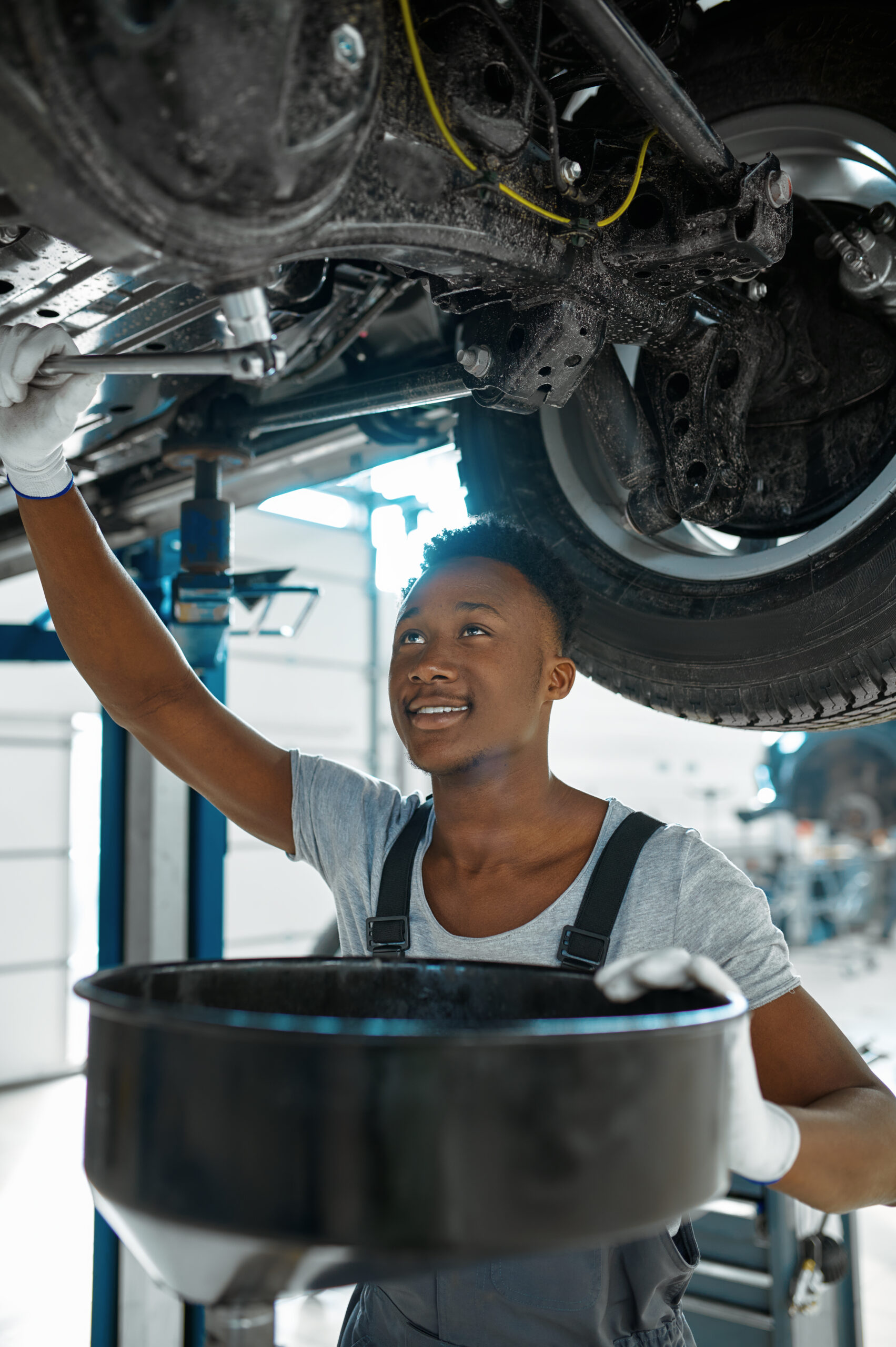 A male mechanic, wearing coveralls and safety glasses, carefully drains used motor oil from a car's engine into a collection pan.