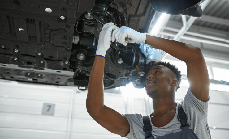 A male mechanic, wearing a blue jumpsuit and safety glasses, tightens a bolt on a car's suspension system while the car is lifted on a hoist.