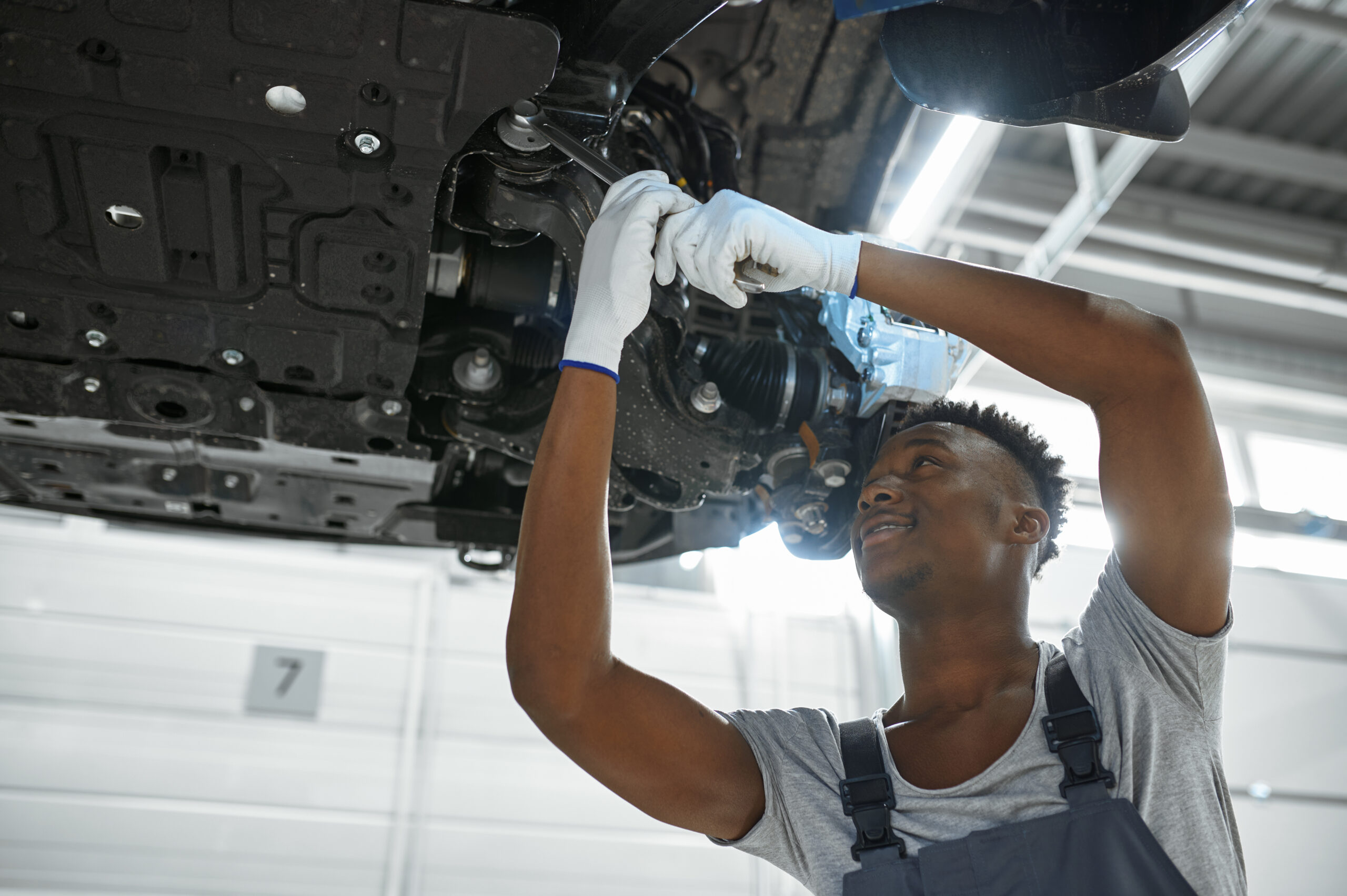 A male mechanic, wearing a blue jumpsuit and safety glasses, tightens a bolt on a car's suspension system while the car is lifted on a hoist.