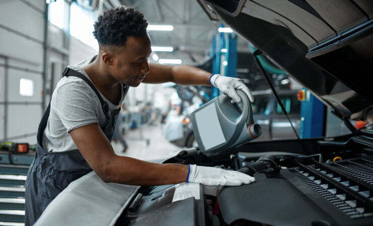 A male mechanic, wearing a blue jumpsuit and gloves, carefully drains used motor oil from a car engine into a collection pan.