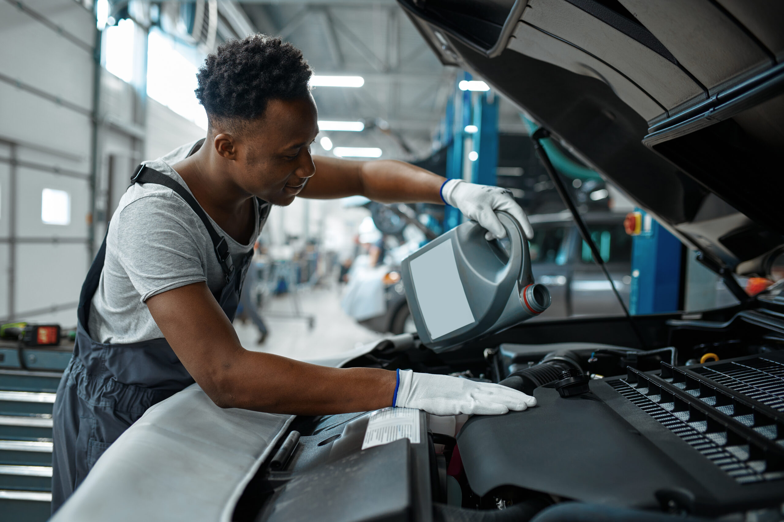 A male mechanic, wearing a blue jumpsuit and gloves, carefully drains used motor oil from a car engine into a collection pan.