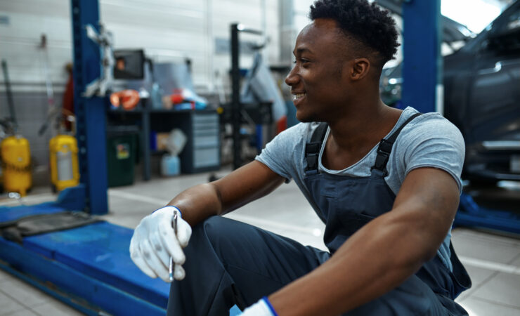 A male mechanic, wearing a blue jumpsuit and holding a wrench, sits on a car lift in a garage, looking up at the undercarriage of a car.