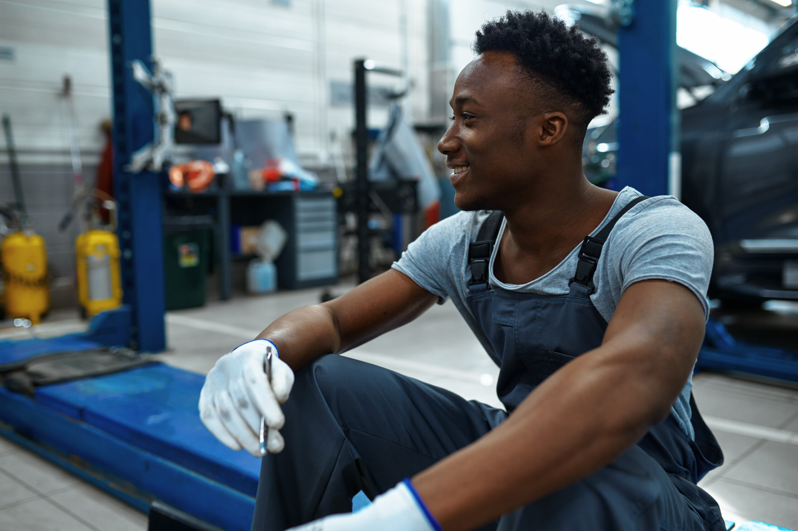 A male mechanic, wearing a blue jumpsuit and holding a wrench, sits on a car lift in a garage, looking up at the undercarriage of a car.