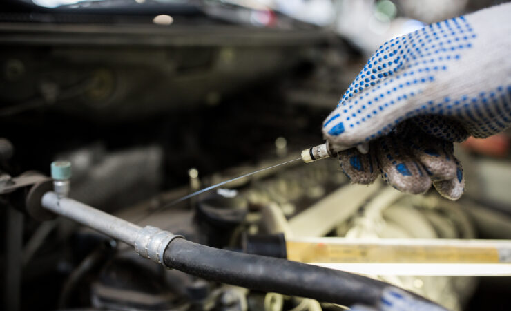 A mechanic, wearing a blue jumpsuit and gloves, holds a dipstick and examines the oil level on it.