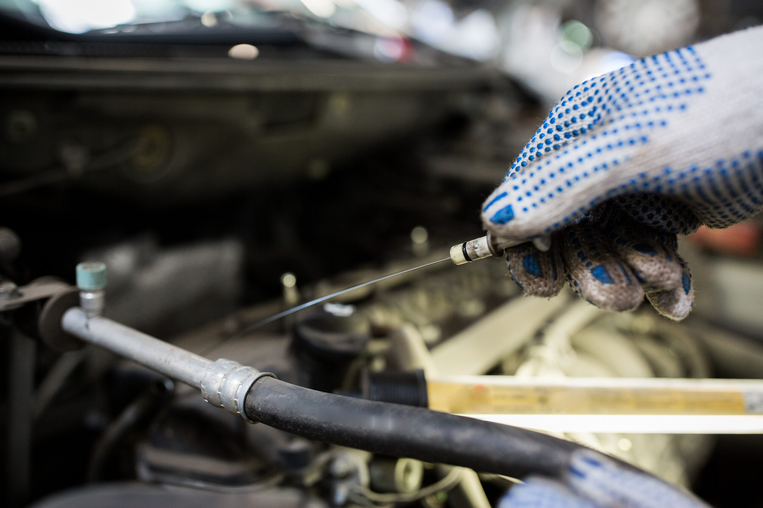 A mechanic, wearing a blue jumpsuit and gloves, holds a dipstick and examines the oil level on it.