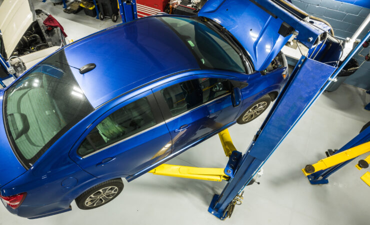 A modern compact car raised on a lift in a brightly lit service bay, with a mechanic inspecting its undercarriage.